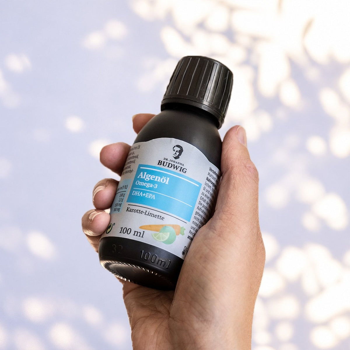 Hand hält eine Flasche Dr. Johanna Budwig Algenöl Omega-3 Karotte-Limette vor einer sonnigen, fliederfarbenen Wand